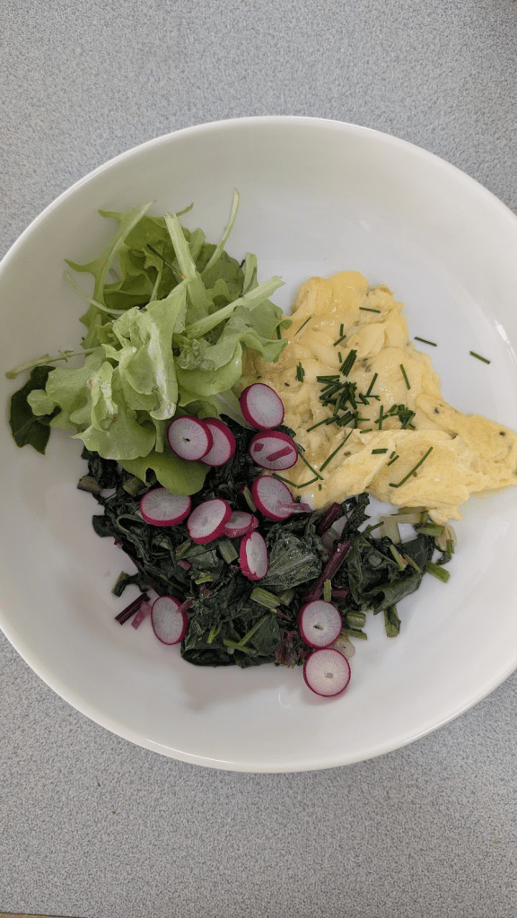 A bowl containing sautéed beet greens with sliced radishes, fresh mixed salad leaves, and an omelet, placed on a gray countertop.