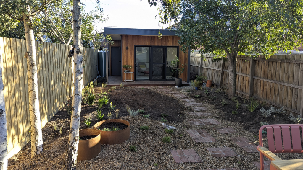 A small urban garden featuring freshly prepared soil ready for planting, with a rustic wooden structure in the background, surrounded by trees and a wooden fence.
