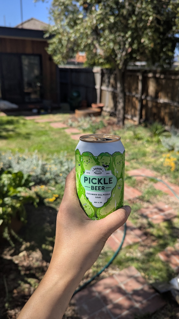 A hand holding a can of 'Pickle Beer' in a garden setting with greenery and brick pathways in the background.
