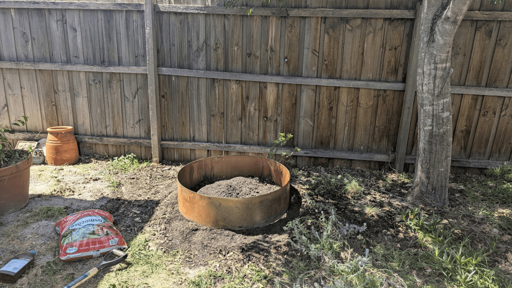 A garden corner being prepared for planting, featuring a metal landscape ring filled with soil, surrounded by plants, a terracotta pot, and gardening tools on the ground.
