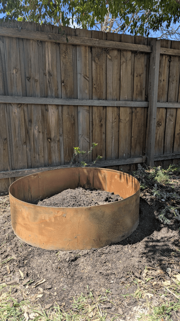 A metal garden bed installed in a sunny corner of a garden, surrounded by freshly turned soil and a wooden fence in the background.