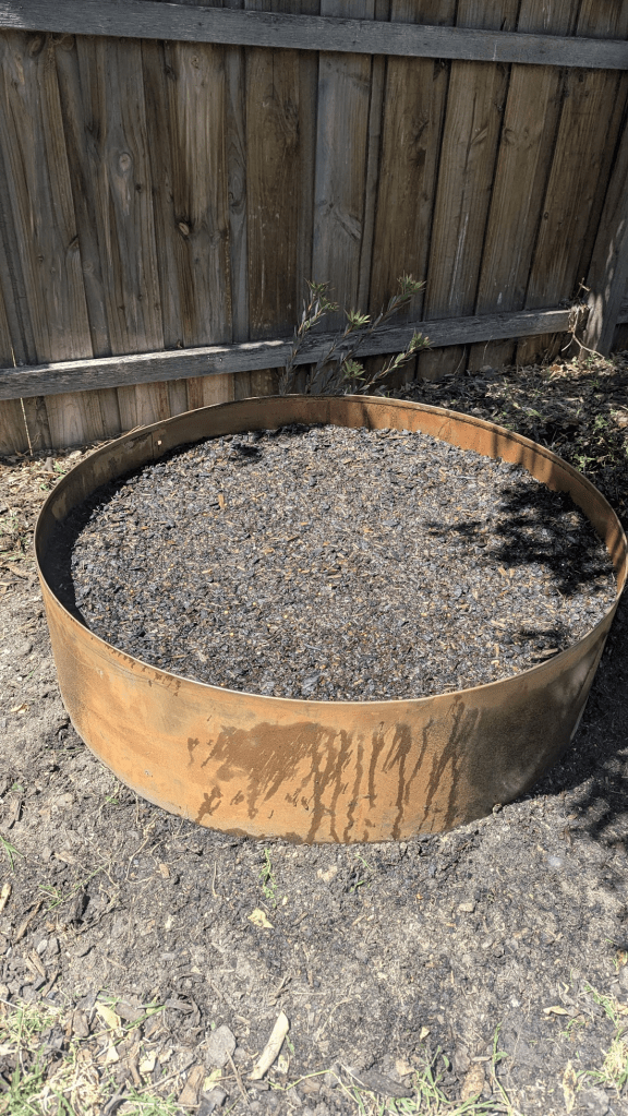 A circular metal garden bed filled with dark, damp soil, positioned against a wooden fence in a backyard.